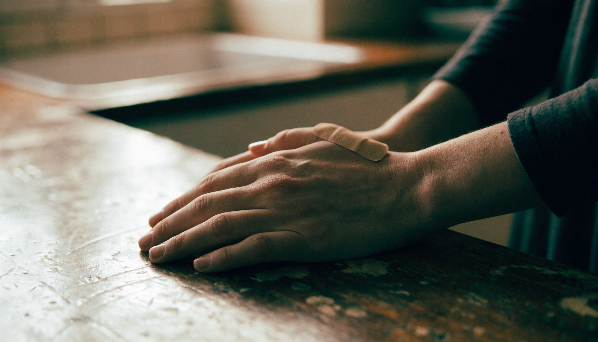 An adult's hands resting on a worn wooden kitchen counter, a simple fabric bandaid on one thumb, in soft morning light.