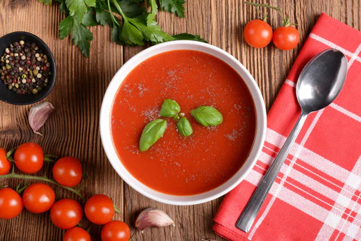 tomato soup with basil garnish in a white bowl surrounded by garlic pepper tomatoes parsley spoon and napkin
